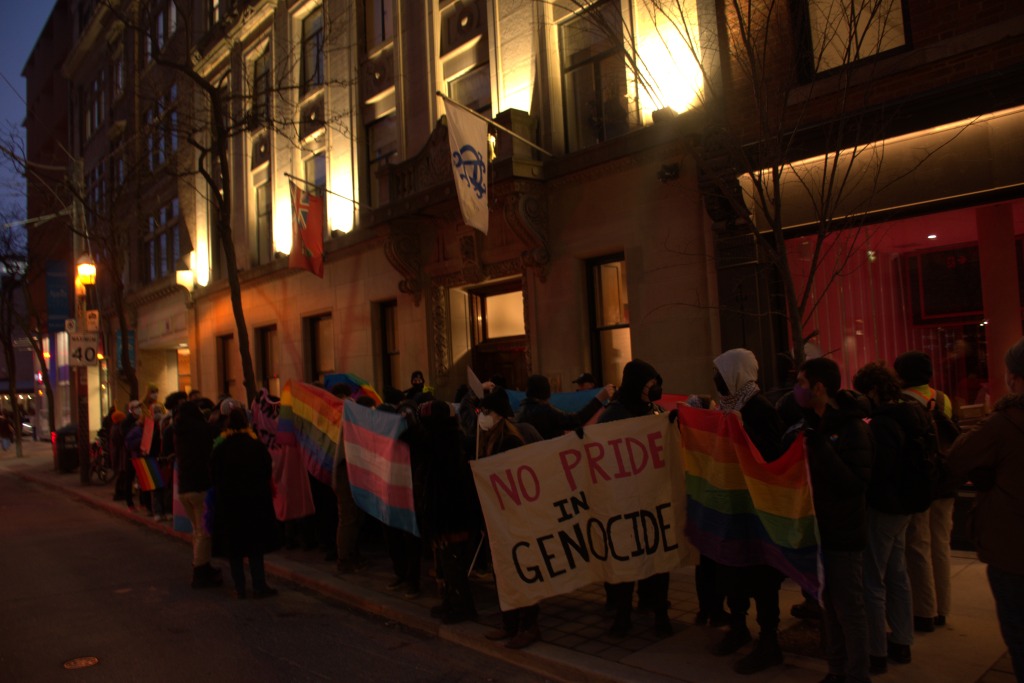 Student protesters on a sidewalk outside of a white marble buulding. Protesters are holding up pride falgs and a banner which reads "No pride in genocide." The Ontario provicial flag and Albany Club flag hang off the building above the protesters.